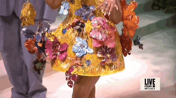 Met Gala 2024 gif. Closeup of the top of Nicki Minaj's yellow Marni mini dress with a low-cut v-neckline and 3D multicolored metal flowers that seem to sprout all around her. The dress flares out in a stiff structured pattern from her waist and comes to about her mid thigh.