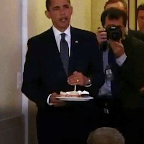 Political gif. Barack Obama walks in, his posse close behind. He carries a plate of cupcakes, cupping his hand to protect the flame and singing “happy birthday to you!”