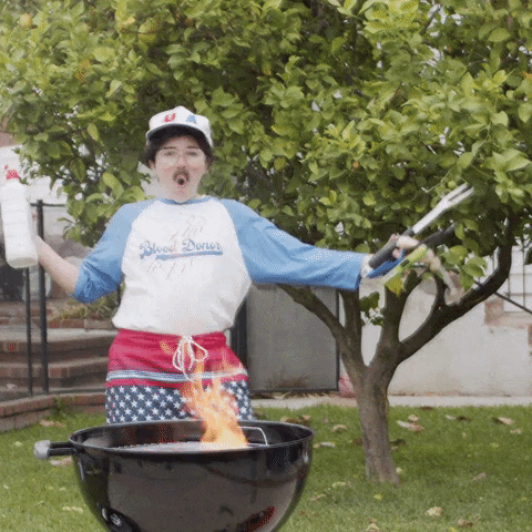 Video gif. Natalie Palamides wears a baseball shirt, fake mustache, and patriotic shorts standing in front of a flaming charcoal barbecue grill in a backyard setting. She's holding BBQ tools and lighters in one hand and lighter fluid in the other, leaning back and thrusting her hips. Text, "Happy Father's Day."