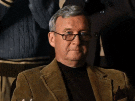 Video gif. Three distinguished looking men clap slowly behind a man sitting in a chair. The man sitting holds a glass of alcohol up and nods proudly.