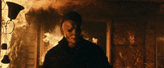 Movie gif. James Jude Courtney as Michael Myers in "Halloween Kills," walks toward us, away from a fire that rages out of a doorway behind him.