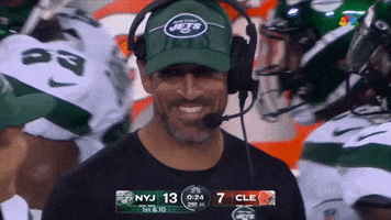 Sports gif. Aaron Rodgers wears a headset and smiles as he talks to another player, both wearing green Jets trucker hats. 