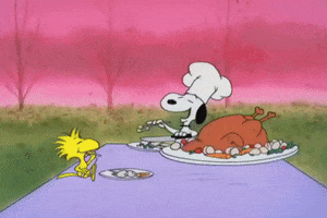 Peanuts gif. Wearing a white chef's hat, Snoopy sits and eats with Woodstock at a table holding an enormous turkey.