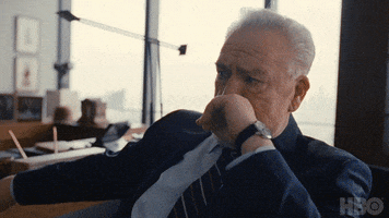 TV gif. Brian Cox as Logan Roy on Succession angrily leaps up from his seat and is held back by an office full of people. 