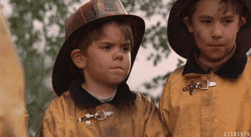 Movie gif. Travis Tedford as Spanky from The Little Rascals is dressed in a tiny, ash-covered firefighter's uniform. He gives us a playful wave as if saying "bye!"
