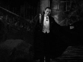 Movie gif. Bela Lugosi as Count Dracula in the 1931 Dracula stands in front of a grand, haunting staircase and spreads his cloaked arms out, saying, "I bid you welcome," which appears as text. The only source of light comes from the candle he holds, and a creepy smile plays upon his face.