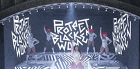 SNL gif. Megan Thee Stallion is on stage at SNL, raising her microphone in the air as she stands in front of a backdrop that says, "Protect Black Women." Four background dancers stand on platforms and do the same pose, all wearing bright red wigs and black and white spiral leotards. 