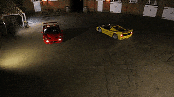 Video gif. Two red and yellow Ferrari F50s face off doing burnouts in an empty lot at night.