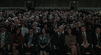 Movie gif. We see a large audience start applauding enthusiastically. We move up higher as balloons float up from the crowd, people stand, and acrobats come down from the ceiling.