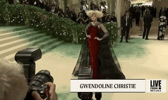 Met Gala 2024 gif. Gwendoline Christie wearing a red Maison Margiela gown and bold, oversized blonde wig, fluffs her sheer black cape, maintaining her posture as she slips into a statuesque pose.