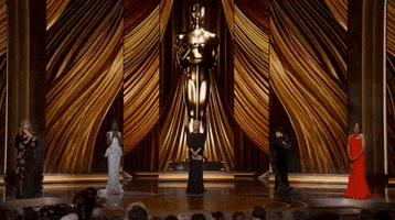 Oscars 2024 gif. From left to right, Mary Steenburgen, Lupita Nyong'o, Jamie Lee Curtis, Rita Moreno and Regina King stand in a horizontal line on the golden stage with a giant Oscars statue towering behind them. Steenburgen and Nyong'o clap.