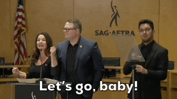 Video gif. Fran Drescher speaks with conviction into a microphone as a man in a blue suit jacket nods behind her in agreement and another signs what she is saying. Text, "Let's go, baby!" Behind them is the SAG-AFTRA logo and an American flag.