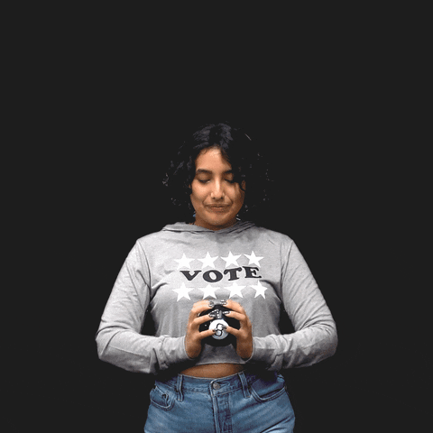 Video gif. Woman wearing a shirt with the word “Voter” shakes a Magic 8 ball in front of herself against a black background. Gold stars appear around her as she reads the prediction. She smiles, and the prediction appears above her head in a blue triangle with the message, “I will be vote ready.”