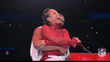 Celebrity gif. Alicia Keys and Usher embrace on stage at the Super Bowl halftime show. Usher gives Alicia a back hug and she closes her eyes and laughs with her mouth open.