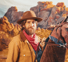 Video gif. Man wearing a cowboy outfit and riding a fake horse smiles and tips his hat in a gentile sort of way against a red rock desert backdrop. 