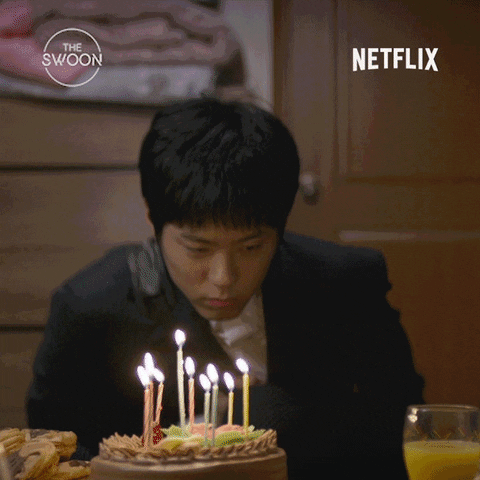 TV gif. An actor in a K-drama Netflix show sits behind a birthday cake with lit candles as someone's arms reach in to put a large birthday hat on his head, as he responds with begrudging compliance. 
