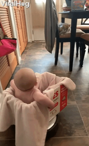 Video gif. An amused, smiling baby sits on a blanket in a Huggies box on top of a Roomba as it glides around the floor.