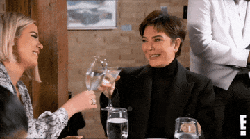 Reality TV gif. Kris Jenner on Keeping up with the Kardashians sits with Khloe Kardashian at a restaurant. Kris holds a martini glass with a big smile and Khloe clinks her glass to toast her mother. 