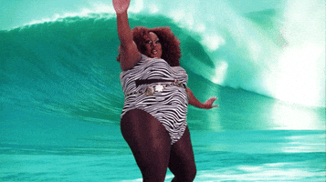 Video gif. A confident fat woman with a big afro reels and regains her balance as she surfs on an artificial surfboard.