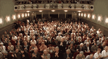 Video gif. In a packed theater, a cheering crowd gives a standing ovation.