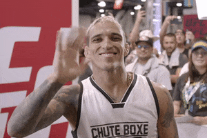 Sports gif. Charles Oliveira from UFC smiles cheekily at the camera while waving and squeezing his fingers cutely.