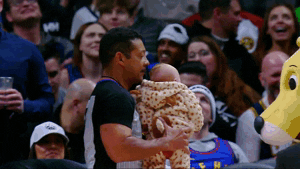 Sports gif. Baby wearing a leopard print onesie is held up like Simba in the Lion King by a referee in the middle of an NBA game as fans cheer in the bleachers behind him.