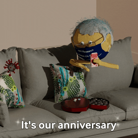 Digital art gif. A man wearing a mask offers a spinning COVID cell a box of medicine. They both sit on a couch and the text below reads, "It's our anniversary."