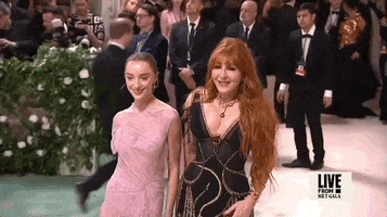 Met Gala 2024 gif. Snap-zoom on Phoebe Dynevor wearing an ethereal Victoria Beckham gown, poses for the cameras holding hands with Charlotte Tilbury, who adjusts with a cock of her head.