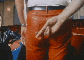 Video gif. Closeup of a hand with fingers crossed held behind the back of a person wearing red leather pants.