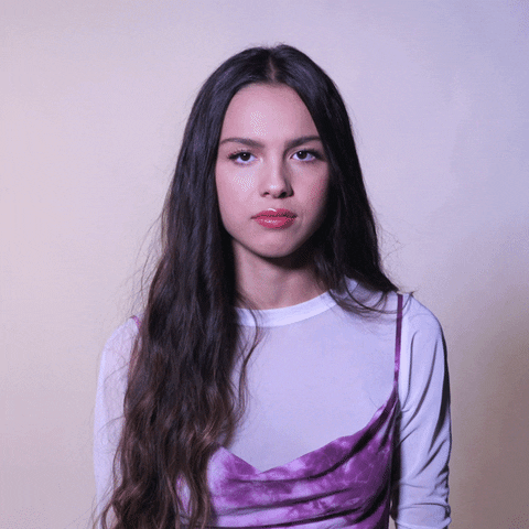 Celebrity gif. Olivia Rodrigo looks at us and crosses her arms together. She contorts her face, making duck lips and scrunching her nose as she thinks. 