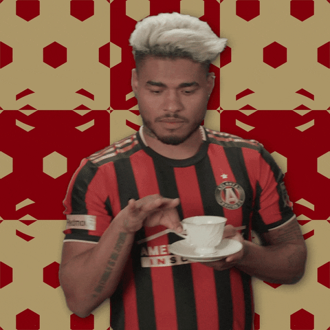 Sports gif. Josef Martínez in his soccer jersey delicately lifts a teacup from its saucer to his lips and takes a sip with an expression of refined elegance. 