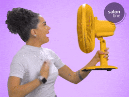 Video gif. Woman holds a big yellow fan to her face and fanning herself while saying, "Que Calor!"