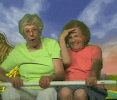 Video gif. Two elderly women are pretending to ride a rollercoaster. They're sitting in front of a green screen of an extreme rollercoaster and are having fun with their faux coaster ride, giggling and putting their hands in the air.