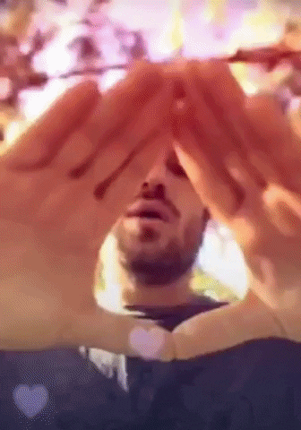 Video gif. Man rotates his hands formed in a triangle towards us and back as he breathes deeply, practicing a reiki healing technique, overlayed with a heart filter.