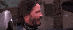 Celebrity gif. Keanu Reeves is in the driver seat of a car and he turns to look at us before giving us a huge wink, moving his entire head as he winks for emphasis.