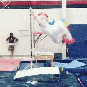 Video gif. Someone dressed in an inflatable unicorn costume swings from a gymnastics bar. They swing all the way around and then flip off of it, and land perfectly on the mat. They jump around excitedly. 