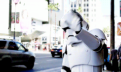 Video gif. Storm Trooper waves at cars in the middle of Hollywood.