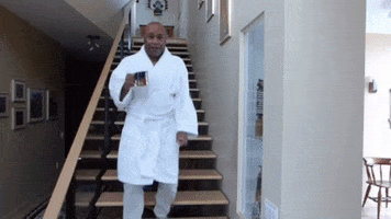 Video gif. A man in a fluffy white bathrobe hustles down a set of stairs before raising his coffee and saying, "Good morning," which appears as text, enthusiastically.