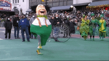 Video gif. Two armless munchkins in costume from Wicked jot through the Macy's Thanksgiving parade.
