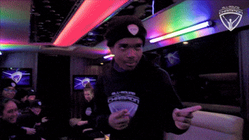 Video gif. Beneath multicolored lights on a low ceiling, a smiling young man makes finger guns to right of frame.