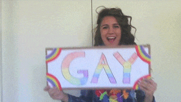 Video gif. A happy woman wearing a lei holds up a sign that reads, "GAY." She looks at the sign and does a fake expression of shock at what she's reading.