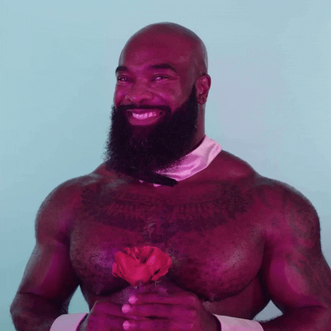 Video gif. Musclebound, shirtless, bald, and bearded man wearing a bowtie and cuffs holds a rose, smiles at us flirtatiously, and says, “Happy hump day.”