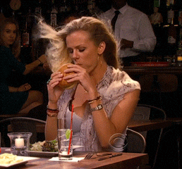TV gif. Brooklyn Decker as Jules in Friends With Better Lives. She's at a dinner table and she's eating a burger in an exaggerated sexy manner as the wind blows her shawl off her shoulders, revealing her tank top.
