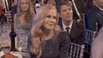 Celebrity gif. Nicole Kidman sits at a table at the 2020 SAG Awards smiles and shrugs her shoulders.