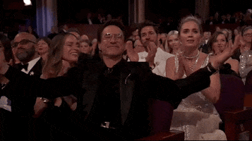 Oscars 2024 GIF. Robert Downey Junior, seated at the Oscars, staggers happily, then claps his hands together decisively, as Emily Blunt looks on, adoring him from behind.