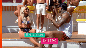 Reality TV gif. Contestants on Love Island are sitting next to each other and the woman says, "The entree is me!" while brushing her hair back. People around her clap and a man next to her gives her a high five.