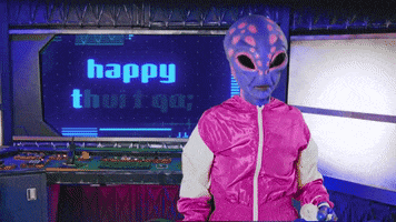 Video gif. A slow zoom in on a person wearing a magenta tracksuit over a purple "gray alien" costume. Their body language seems slightly troubled, and the computer console behind them reads "Happy Tuesday".