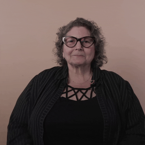 Reaction gif. A Disabled white woman with kinky curly gray hair and big wine-colored cat-eye glasses hugs herself, clutching her heart and relishing in it.