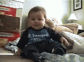 Video gif. Low-angle footage of a baby, sitting on the floor, giggling so hard they gently topple over to one side.