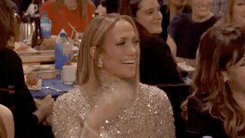 Celebrity gif. At an awards show, Jennifer Lopez smiles and waves from her seat.